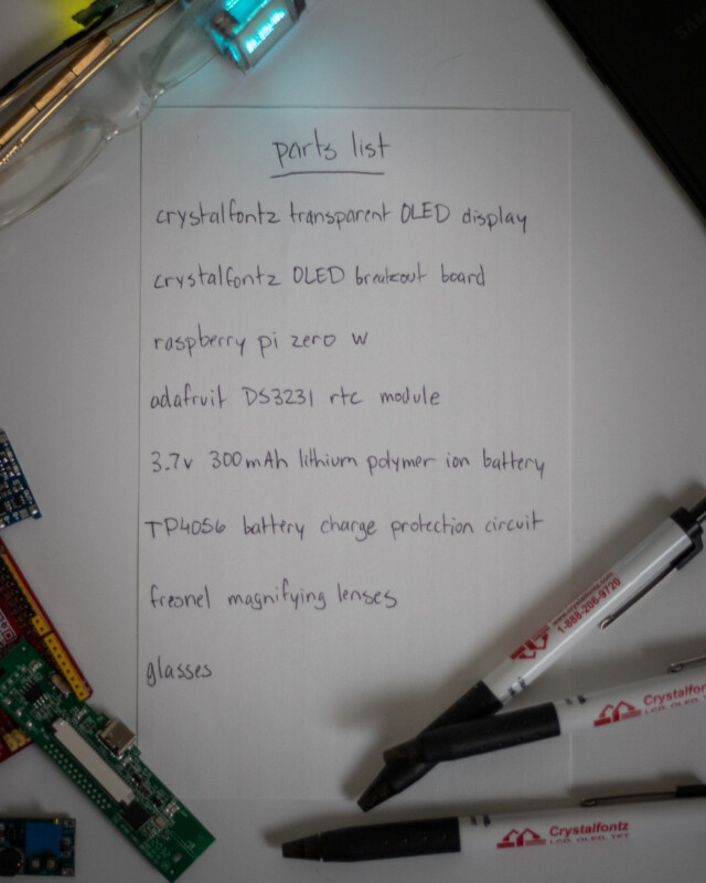 picture of a piece of paper that says: Parts list:
TP4056 battery charge protection circuit
Fresnel magnifying lenses
Glasses
various components and pens are visible on the edge of the paper