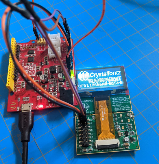 A transparent OLED on a CFA10105 breakout board connected to a Seeedunio using four wires. The OLED is displaying the Crystalfontz logo and the words "Crystalfontz Transparent CFAL12856A0-0151-B"